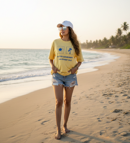 A woman stands on a sandy beach wearing a yellow graphic t-shirt saying better sunburnt than burnt out, denim shorts, and a white cap, with the ocean and palm trees in the background.