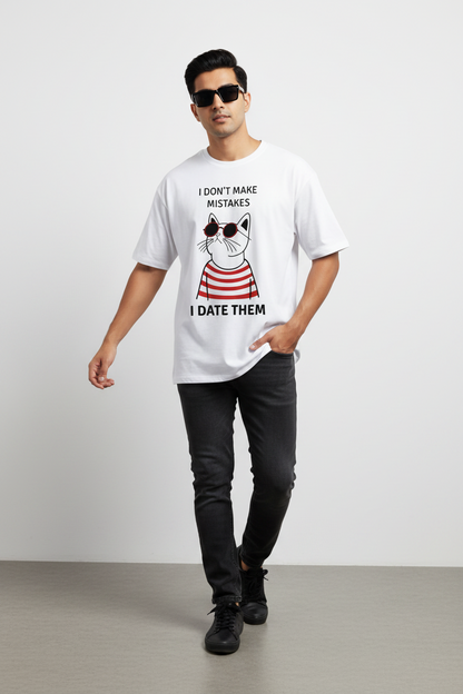 A man modeling a white graphic t-shirt with a cartoon cat wearing sunglasses and the text 'I DON'T MAKE MISTAKES I DATE THEM'. He is also wearing black pants, black sunglasses, and black sneakers, standing against a light gray studio background.