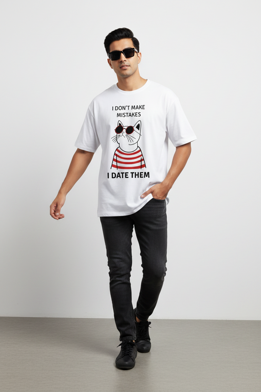 A man modeling a white graphic t-shirt with a cartoon cat wearing sunglasses and the text 'I DON'T MAKE MISTAKES I DATE THEM'. He is also wearing black pants, black sunglasses, and black sneakers, standing against a light gray studio background.