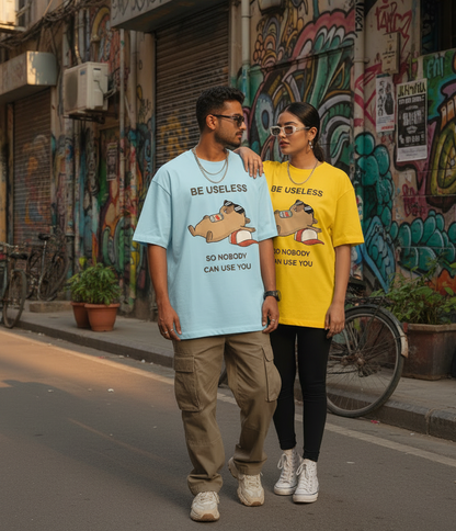 A man and a woman walking side-by-side on a city street with colorful graffiti-covered walls. The man wears a light blue oversized t-shirt with a cartoon graphic and the text 'BE USELESS SO NOBODY CAN USE YOU', while the woman wears a matching yellow oversized t-shirt with the same graphic.