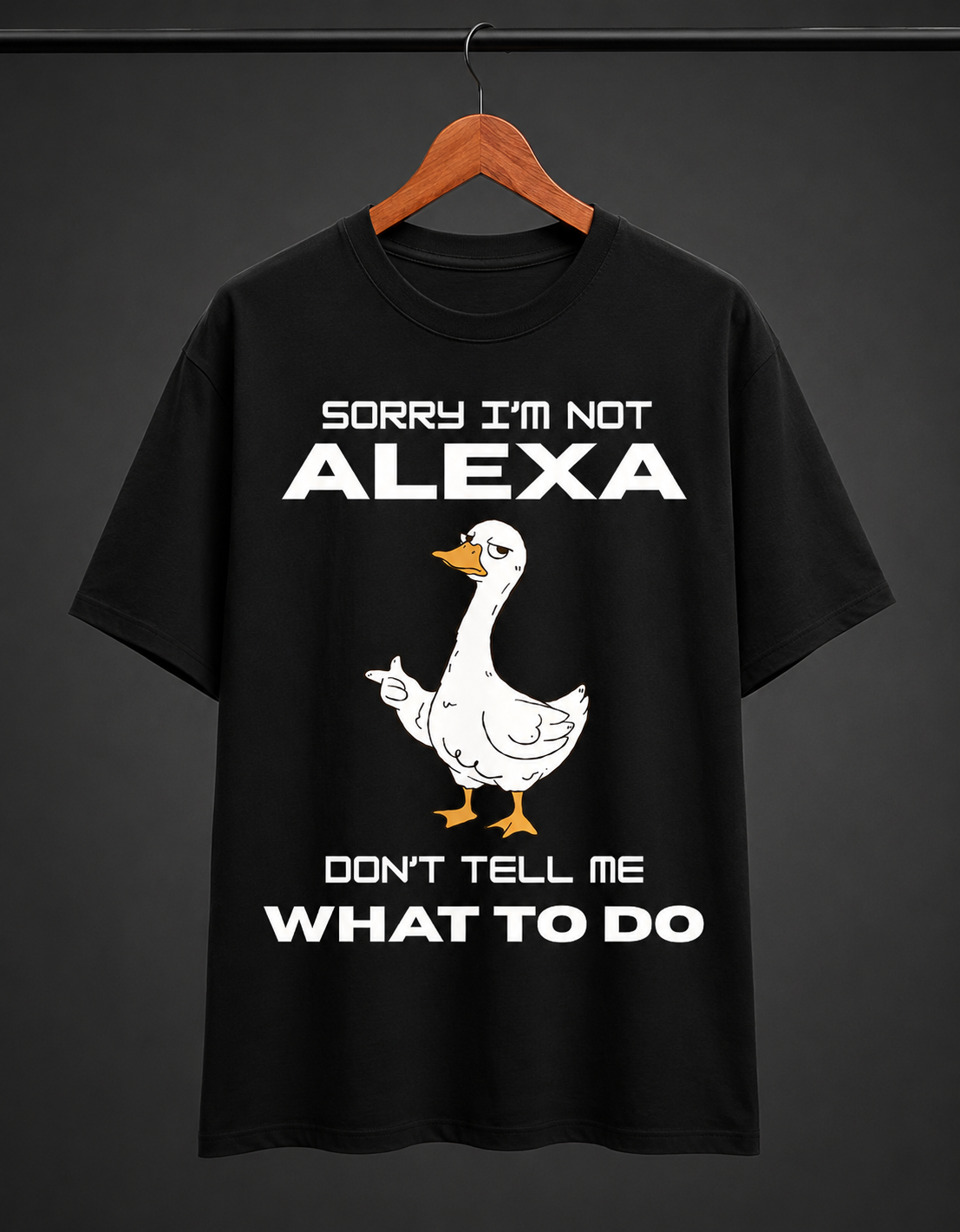 A black short-sleeve t-shirt hanging on a wooden hanger against a dark gray background. The shirt features a white cartoon goose graphic with orange beak and feet, accompanied by the white text 'SORRY I'M NOT ALEXA DON'T TELL ME WHAT TO DO'.